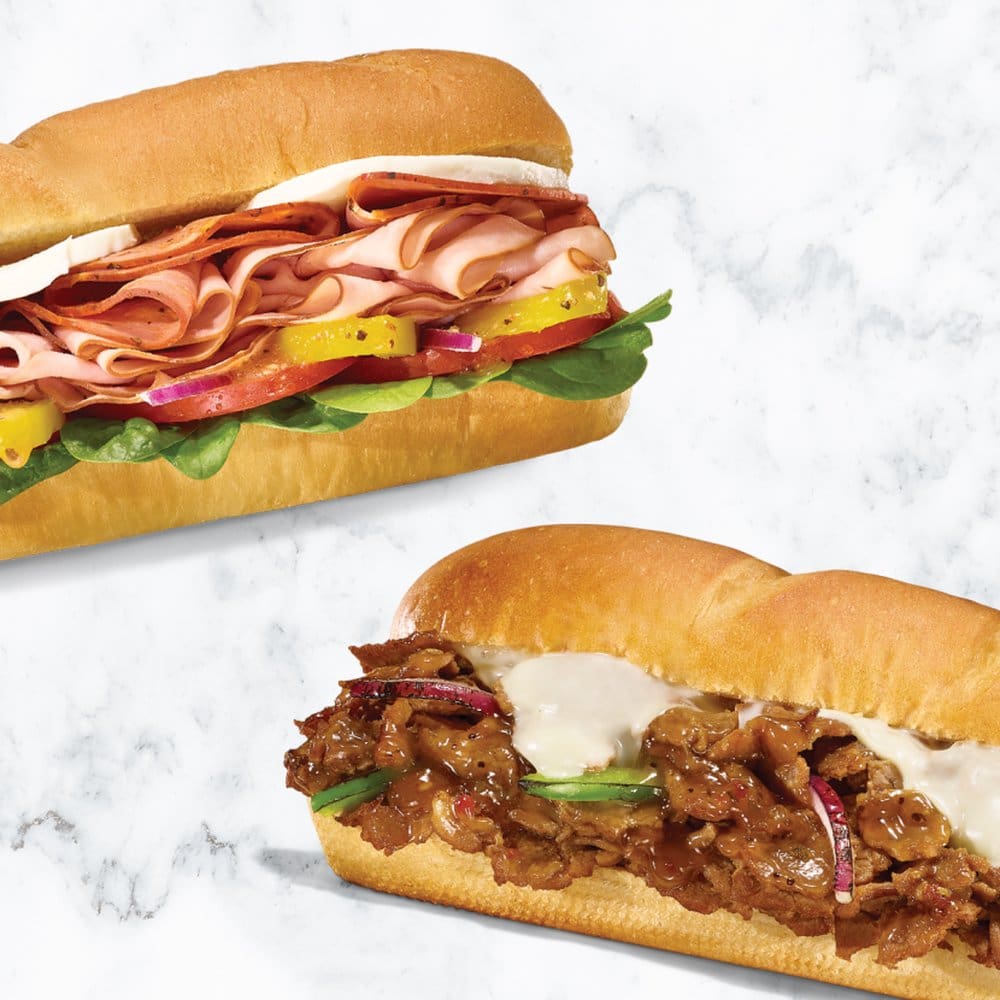 Two submarine sandwiches on a marble surface: one with ham, cheese, peppers, and greens; the other with sliced beef, onions, and sauce.