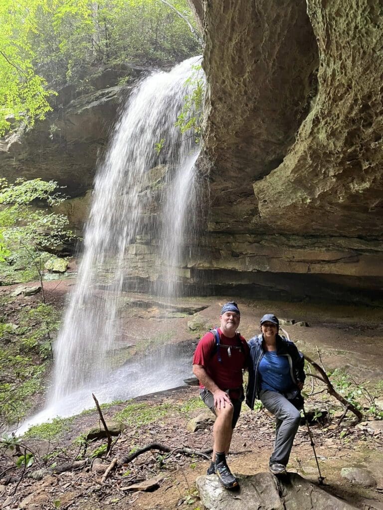 Two hikers pose beside a waterfall in a rocky alcove, with green trees above the scene still bright and lush.