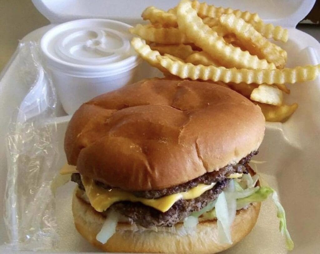 Cheeseburger with lettuce and cheese on a soft bun, crinkle-cut fries, and a cup of white dipping sauce in a takeout tray.