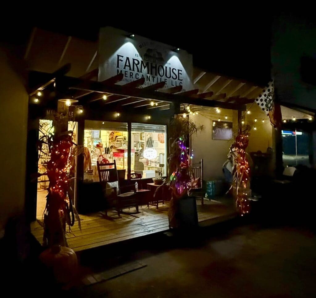 Night exterior of Farmhouse Mercantile storefront with string lights, pumpkins, and holiday decorations on a wooden porch. Inside glow visible through glass doors.