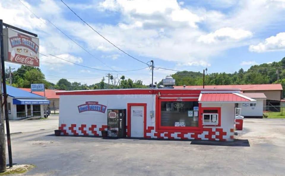 Small, retro-style diner with red trim and a red-and-white checkerboard border, large front window, and a vending machine outside, in a sunny parking lot.