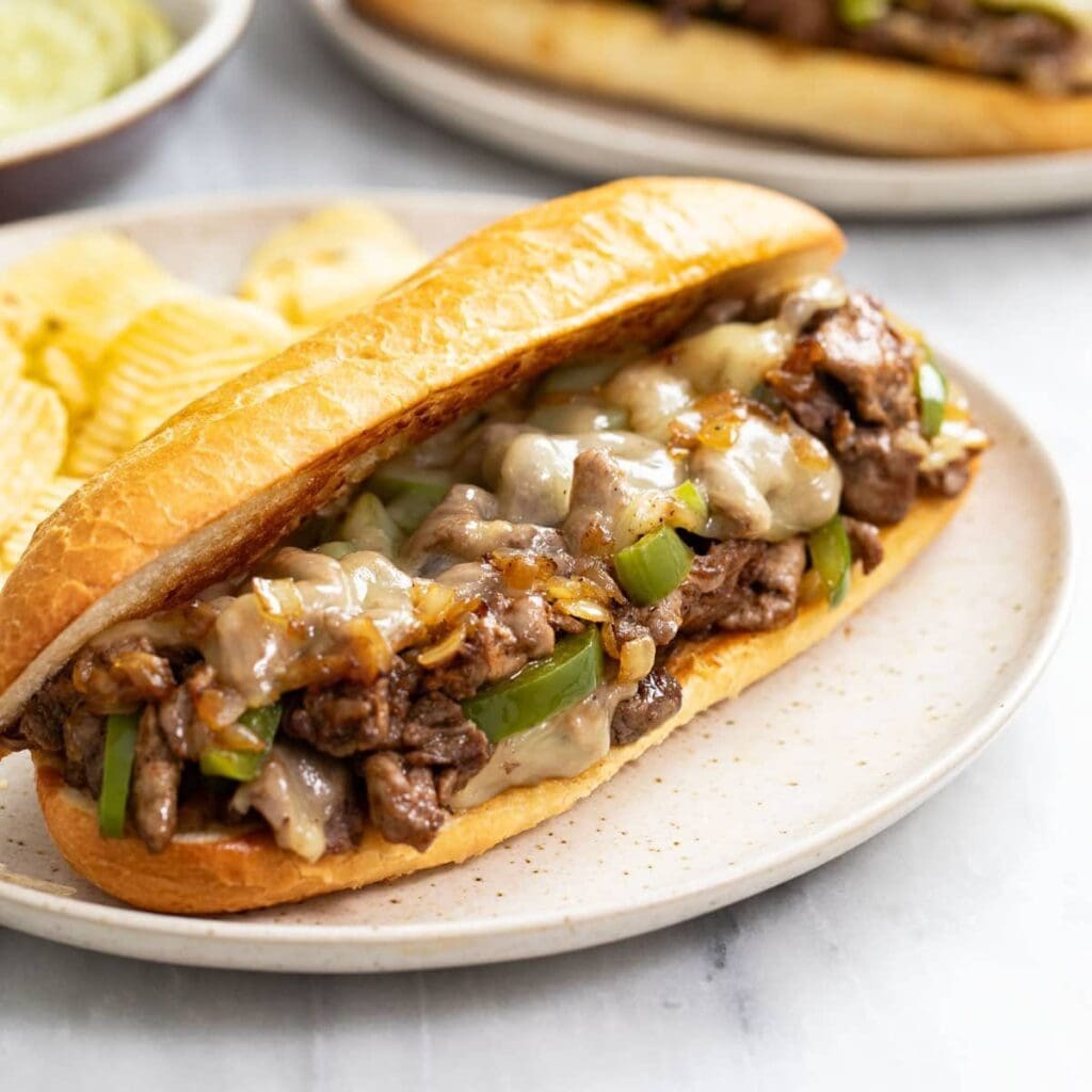 Beef Philly-style cheesesteak on a toasted bun with melted cheese, onions, and green peppers, on a beige plate with chips nearby.