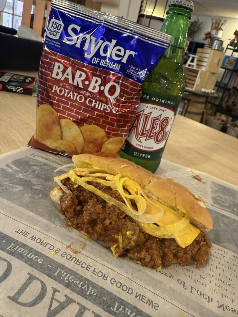 Bag of Snyder's of Berlin BAR-B-Q potato chips and a green bottle sit on a table beside a half-eaten cheeseburger with onions and cheese on a paper-lined surface.