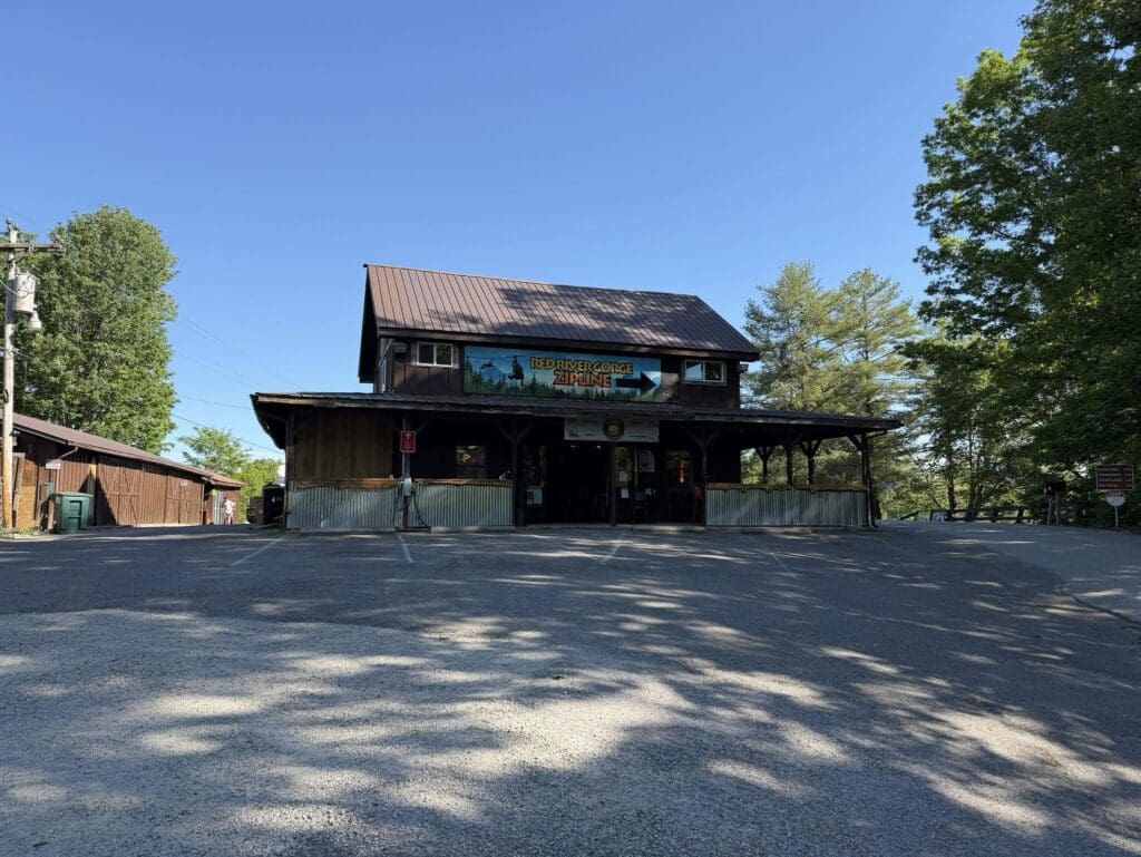 Two-story wooden building with a colorful sign reading 'Red River Gorge Zipline' under a clear blue sky, surrounded by trees.