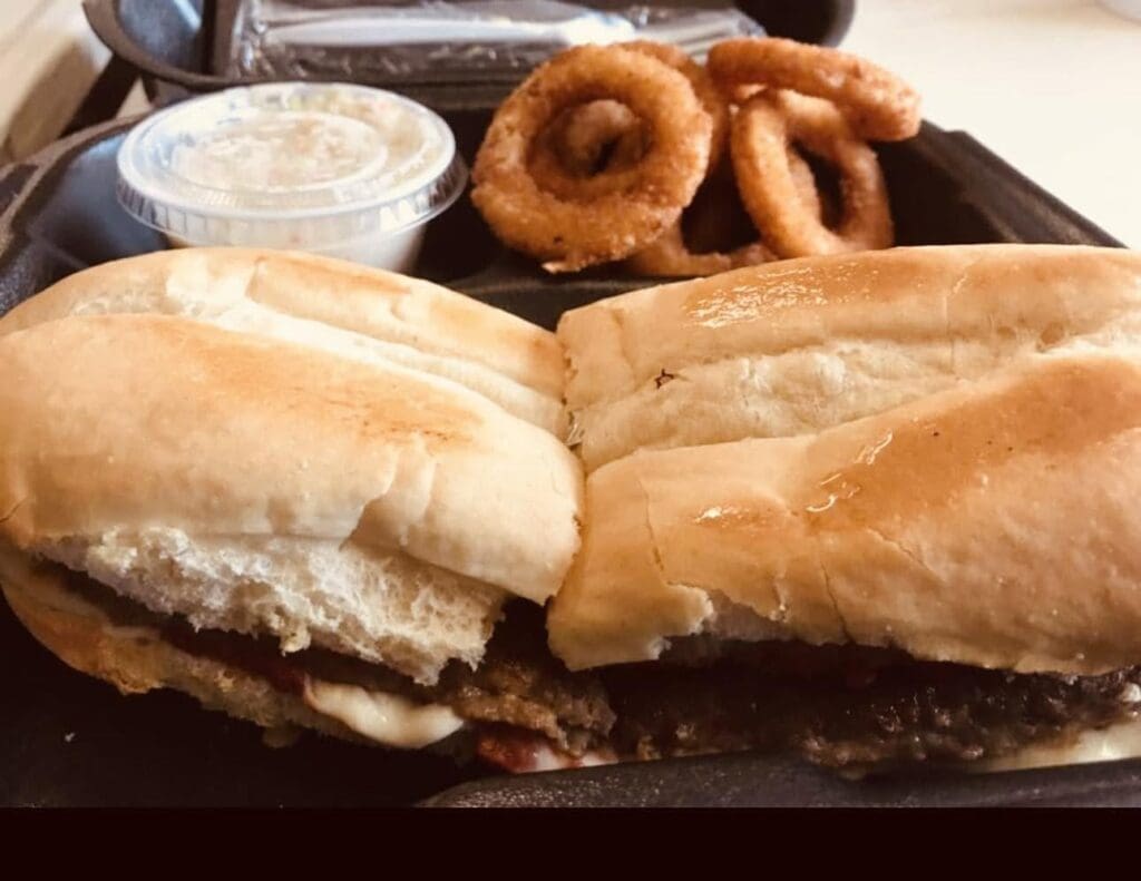 Two hamburger buns with a beef patty and cheese, plus onion rings and a cup of sauce on a black tray.