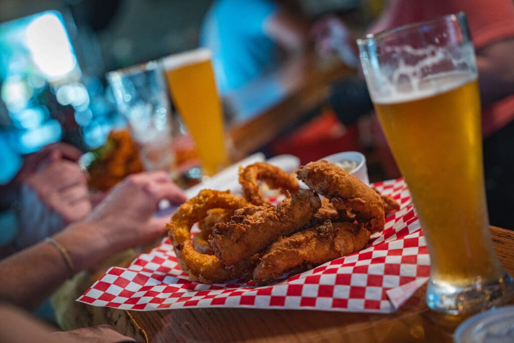 Plate of crispy fried chicken on red-and-white checkered paper, with tall glass of beer in a blurred background.