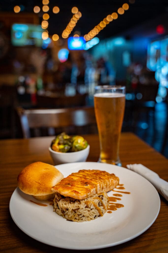 Plate with a bread roll, a glazed fish fillet on rice, and drizzled sauce; beer glass in the blurred background at a restaurant.