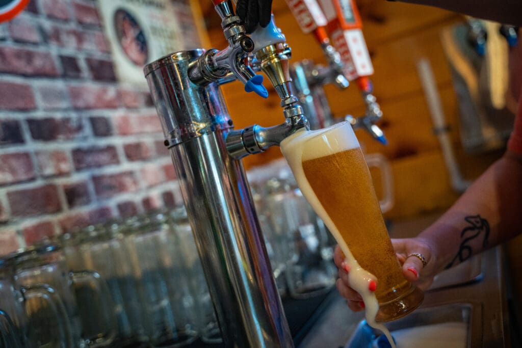 Close-up of a beer tap pouring a foamy lager into a tall glass, with a brick wall background behind the bar.