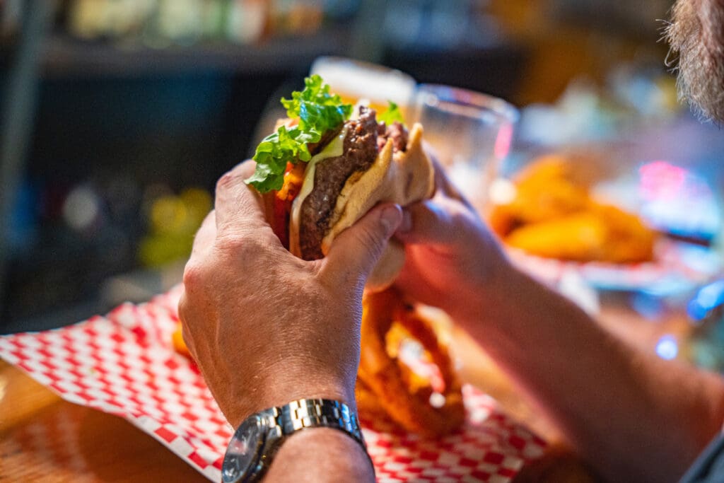 Hands holding a loaded sandwich with lettuce, cheese, and meat over a red-and-white checkered tablecloth with a blurred bar background.