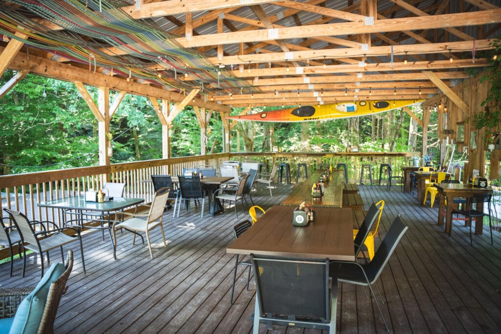 Open-air dining area on a wooden deck with string lights and a colorful kayak hanging from the ceiling, trees in the background.