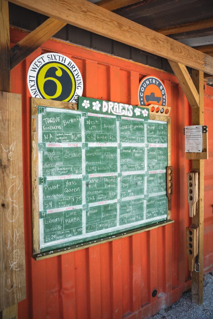 Chalkboard beer menu on a wooden frame mounted to an orange shipping container, with round West Sixth Brewing and Country Boy signs above.