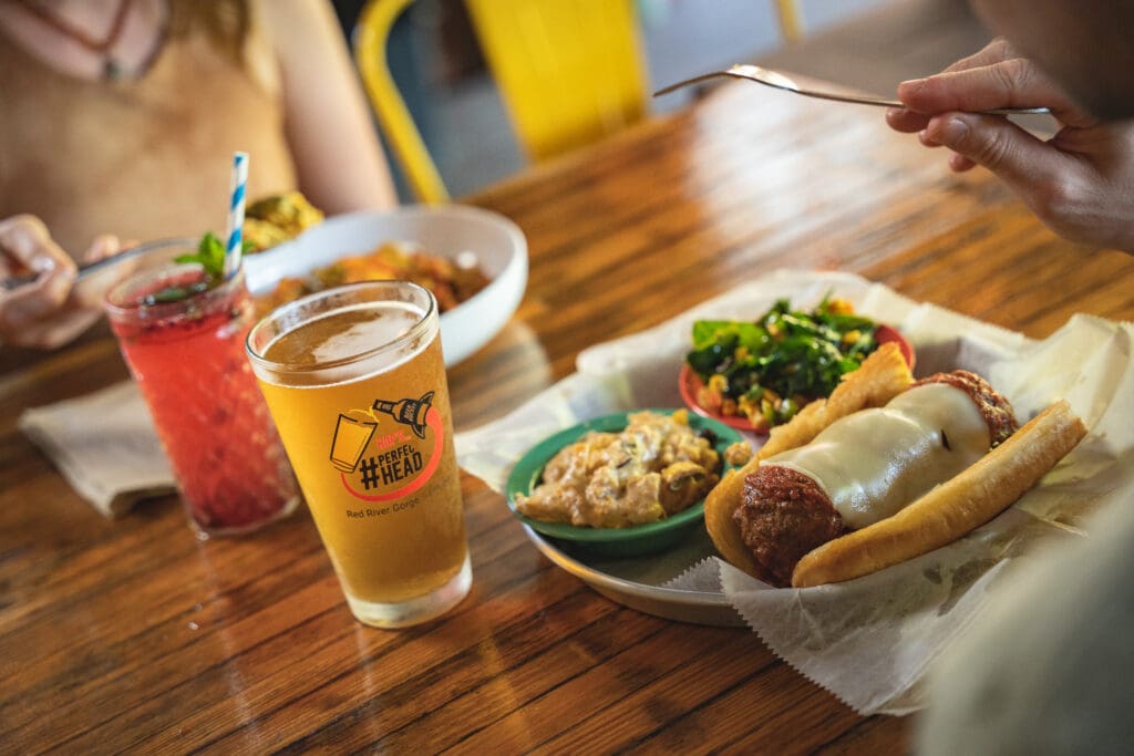 Casual meal scene: hot dogs with toppings on a tray, greens and side dishes, a glass of beer, and a hand holding a spoon over the table.