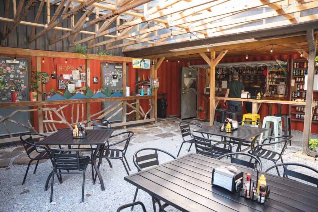 Open-air cafe with black metal tables and chairs under a wooden pergola, string lights, and a red wall interior visible in the background.
