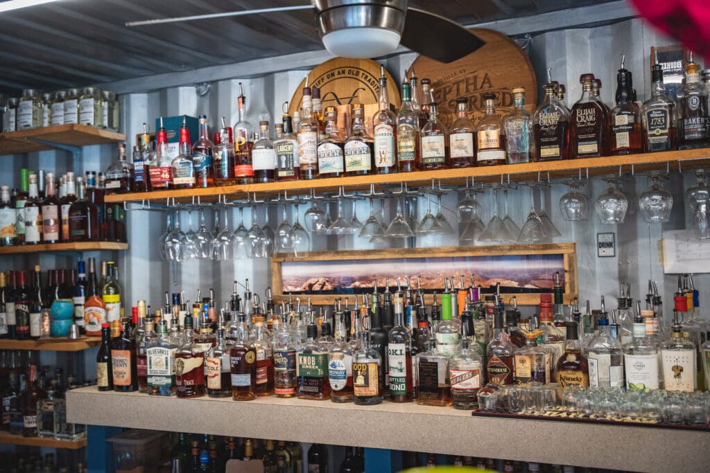 Bar shelf full of assorted whiskey bottles with upside-down wine glasses underneath the counter rail