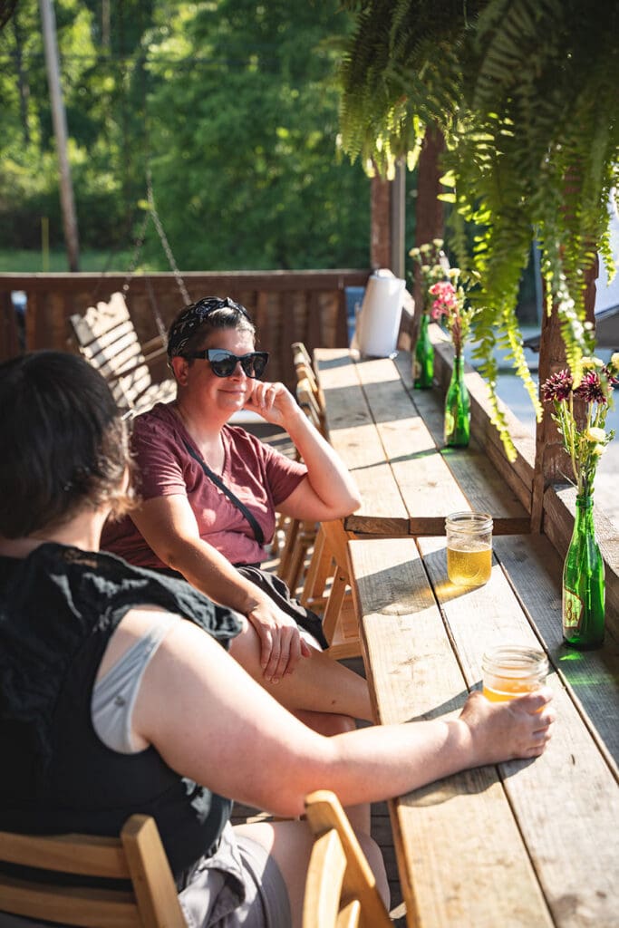 Two friends sit at a sunlit outdoor wooden bar, chatting with beers in jars and green bottles on the table.