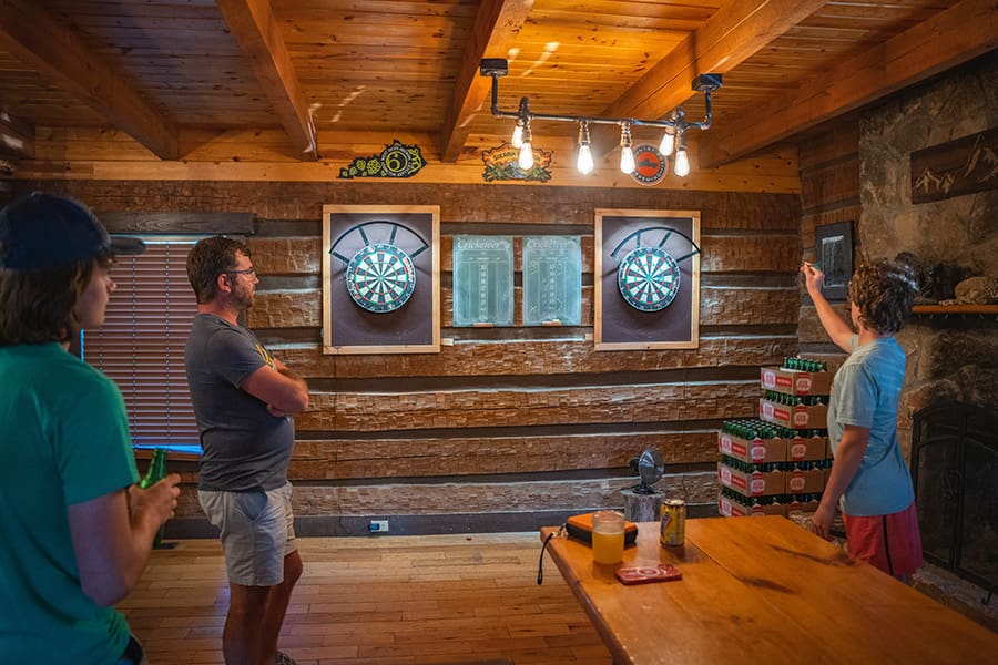 Three friends play darts in a rustic wood-paneled basement with two dartboards and a chalkboard between them.