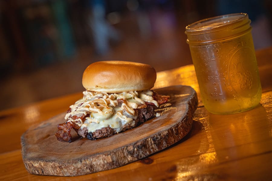Beef burger on a wooden board topped with melted cheese, onions, and bacon, with a mason jar of beer nearby on a wooden table.