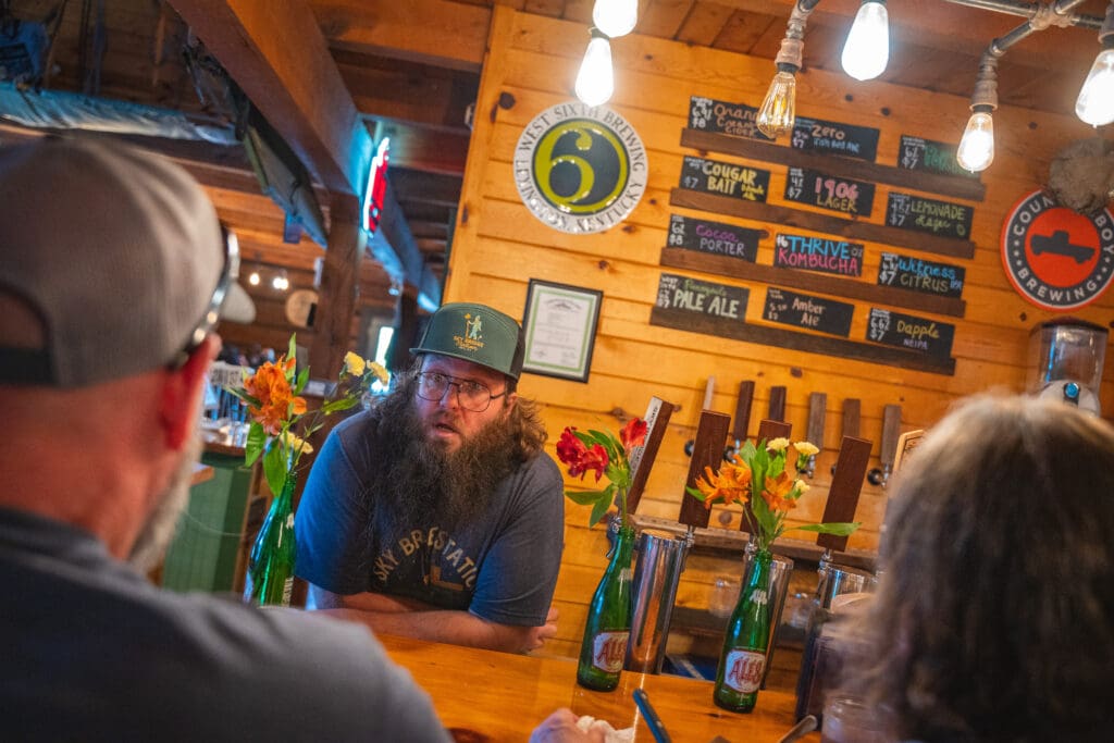 Bearded bartender in a green cap talking to customers at a wooden bar; beer bottles and flowers on the counter.