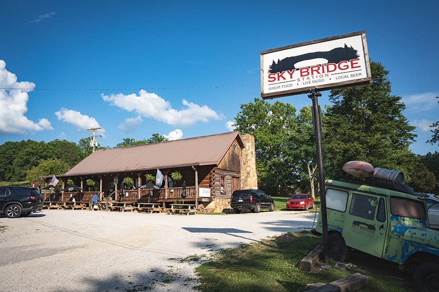 Roadside rustic restaurant with a long wooden porch, a Stone chimney, and a Sky Bridge sign in a gravel lot under a blue sky