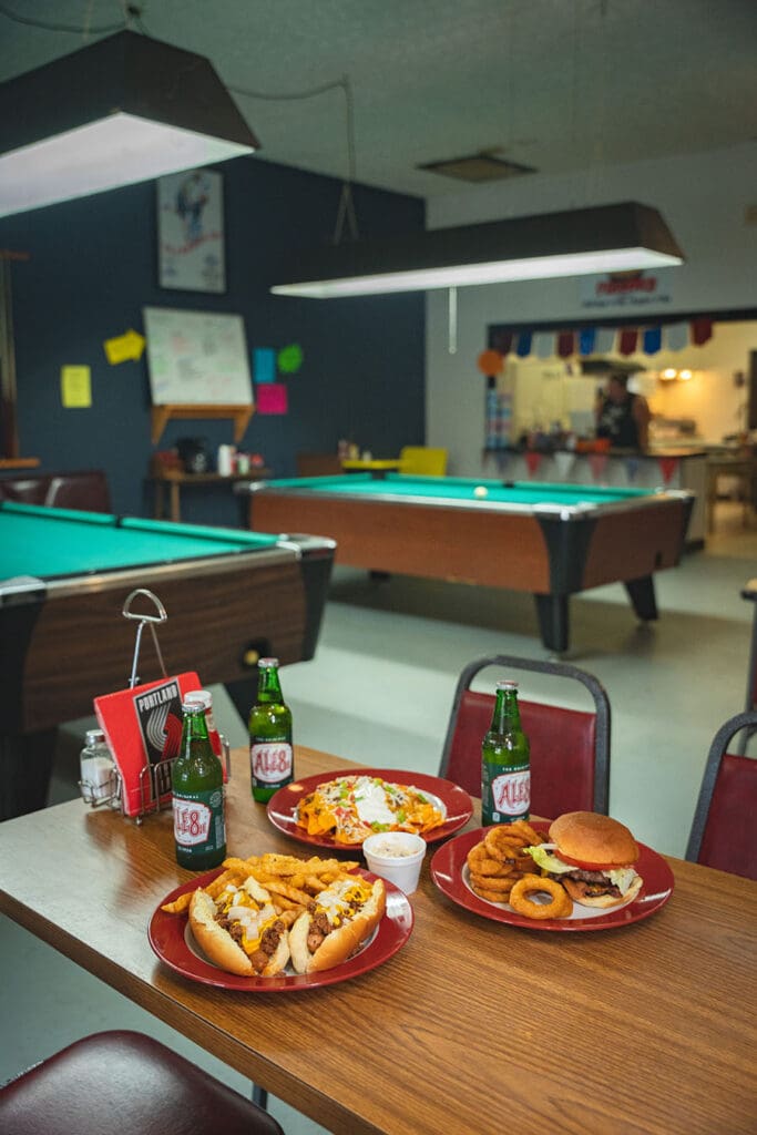 Casual dining setup in a pool hall: three plates of nachos, hot dogs, onion rings, two bottles of Ale8, and a small cup on a wooden table with pool tables in the background.