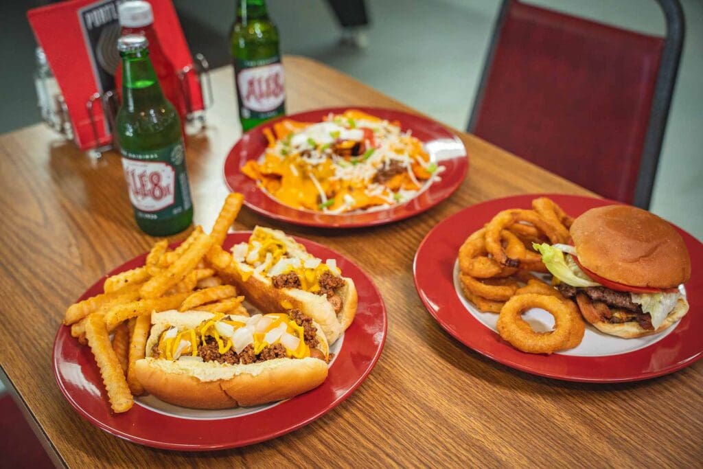 Hot dogs loaded with cheese and onions with fries on a red plate, a burger with onion rings on another red plate, beer bottles on a wooden table nearby.