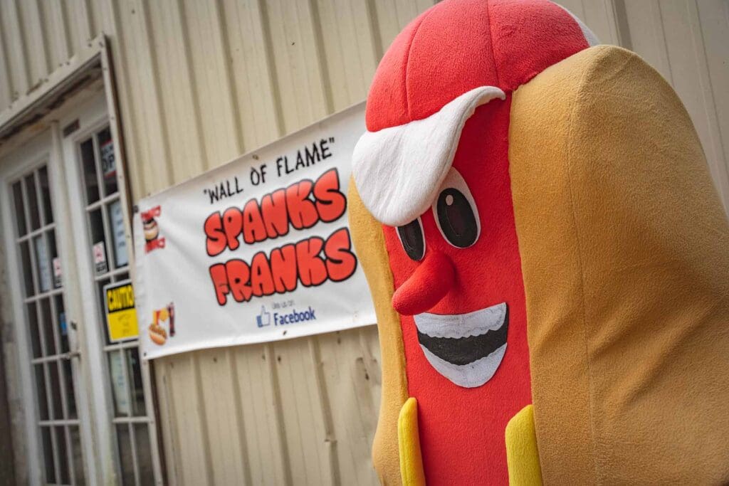 A sausage mascot in a red and tan costume with white gloves stands next to a banner that reads 'Wall of Flame' and 'Spanks Franks' outside a shop.