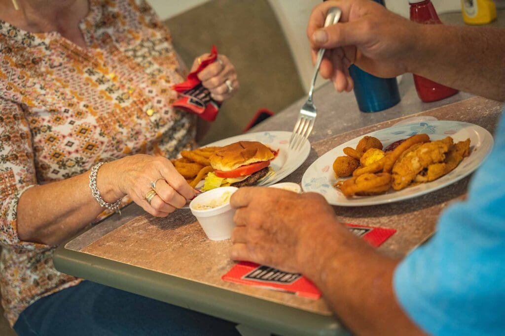 Elderly woman at a counter adds toppings to a hamburger while a man forks a plate of fried snacks nearby.