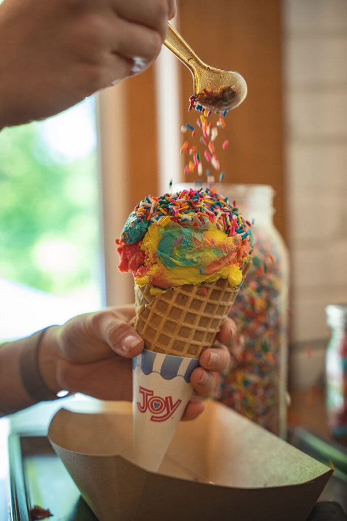 Hand holding a waffle cone with rainbow ice cream as colorful sprinkles fall from a spoon above it.