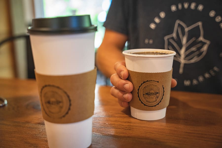 Two disposable cups with brown sleeve on a wooden table; a person holds a smaller cup in the background.