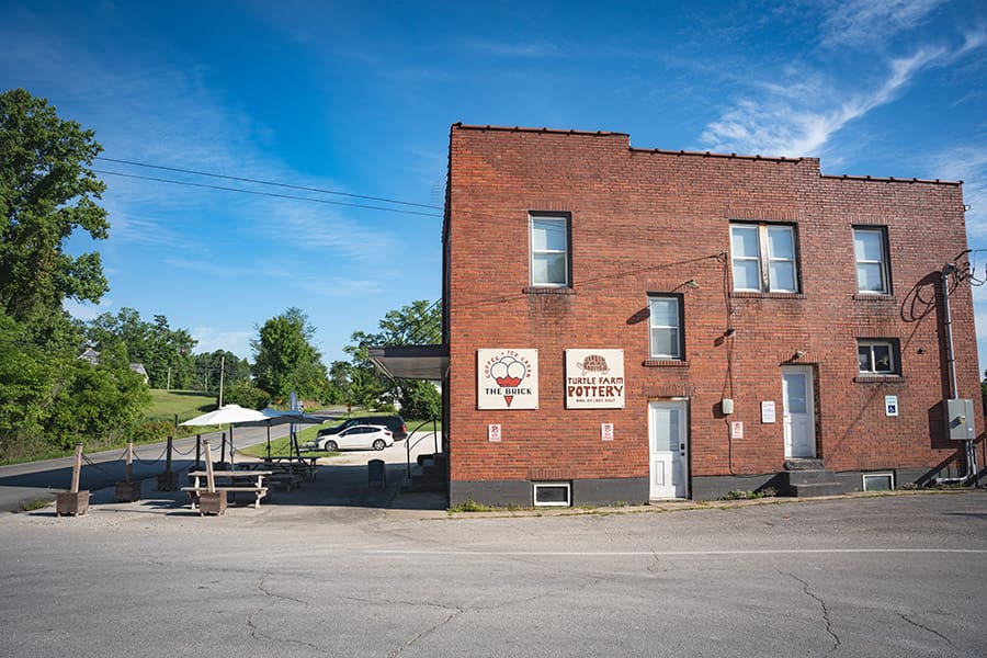 Brick two-story building housing a pottery shop, with signs reading "Turtle Farm Pottery" and "The Brick" on a sunny day, and outdoor seating to the left.