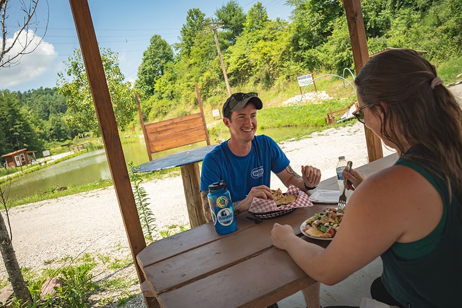 Two friends share a casual outdoor meal at a wooden picnic table beside a small pond, smiling and chatting as they eat and drink.