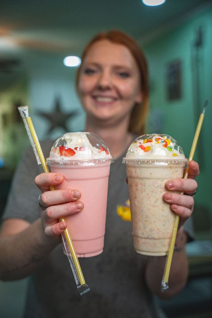 Smiling woman holds two whipped‑cream-topped milkshakes, one pink strawberry and one vanilla with sprinkles, toward the camera.