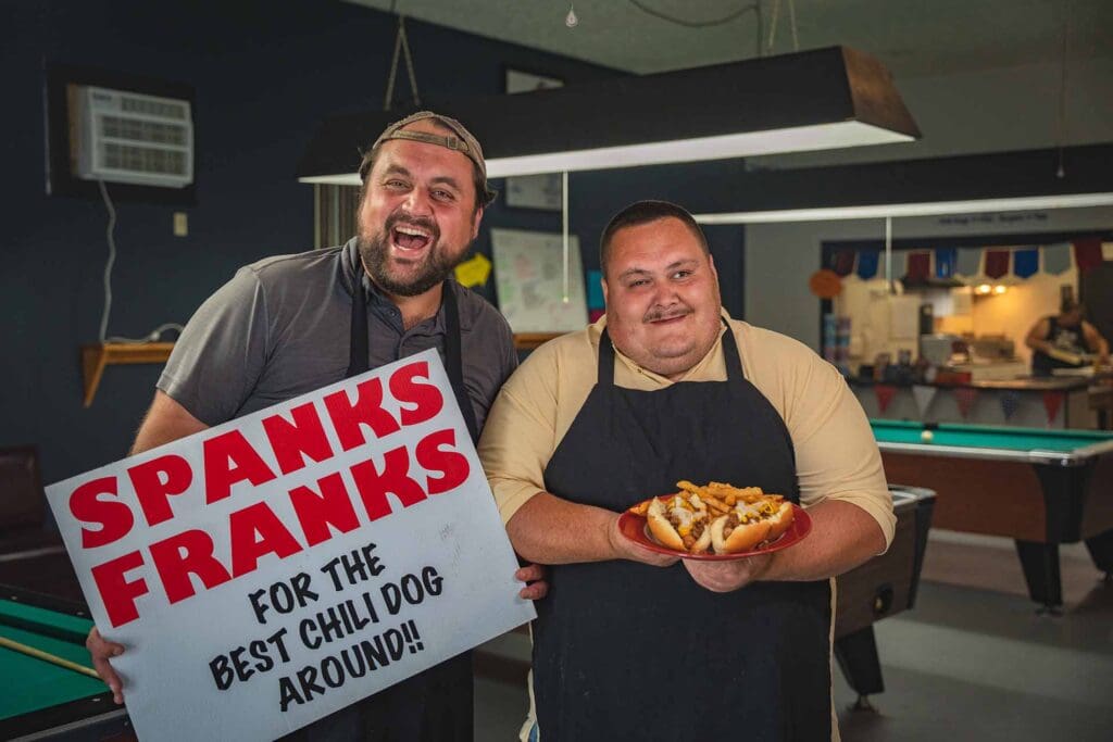 Two men in aprons smiling in a kitchen/diner, one holding a large sign with red lettering while the other presents a plate of loaded hot dogs.