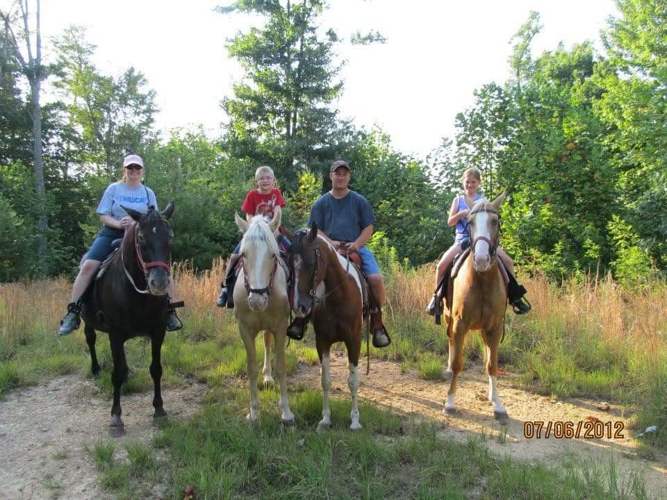 A family of five riding horses on a dirt trail through a grassy clearing with trees in the background.
