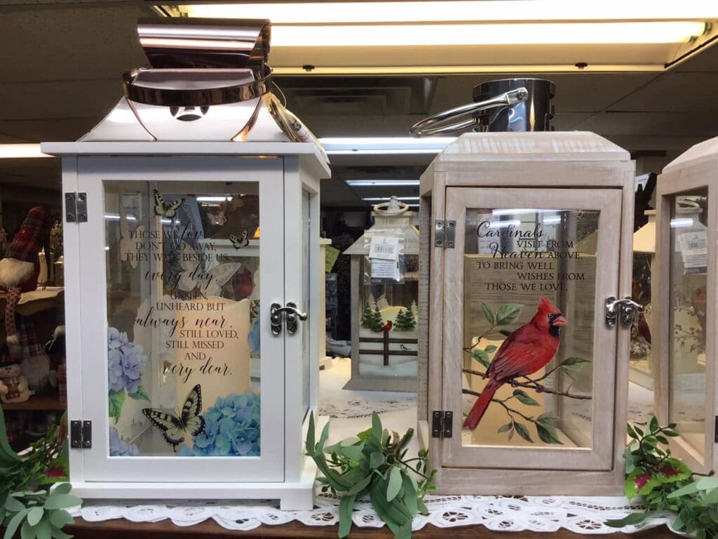 Two decorative glass-front display cabinets side by side on a lace cloth; left shows butterflies and flowers with a sentimental quote, right features a cardinal on a branch with text on the glass.