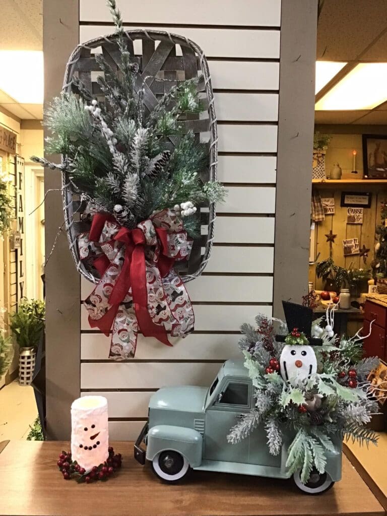 Large hanging Christmas basket filled with frosted pine branches and a red ribbon, mounted on a striped wall behind a shop display.