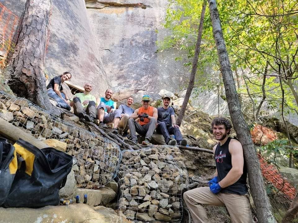 Group of nine men posing on a rocky slope with stacked stones and fencing in a quarry, smiling at the camera.