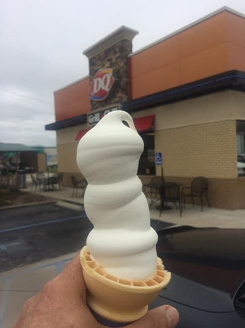 Tall vanilla soft-serve ice cream cone held in hand, with Dairy Queen storefront in the background.