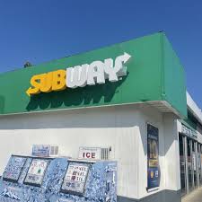 Exterior of a Subway restaurant with a green facade and yellow logo, set against a clear blue sky.
