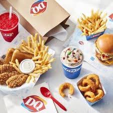 Assorted fast-food spread: fries, chicken tenders with dipping sauce, a burger, onion rings, a soft drink, and a Dairy Queen dessert and cup on a light table surface shaped by fast-food bags and cups.