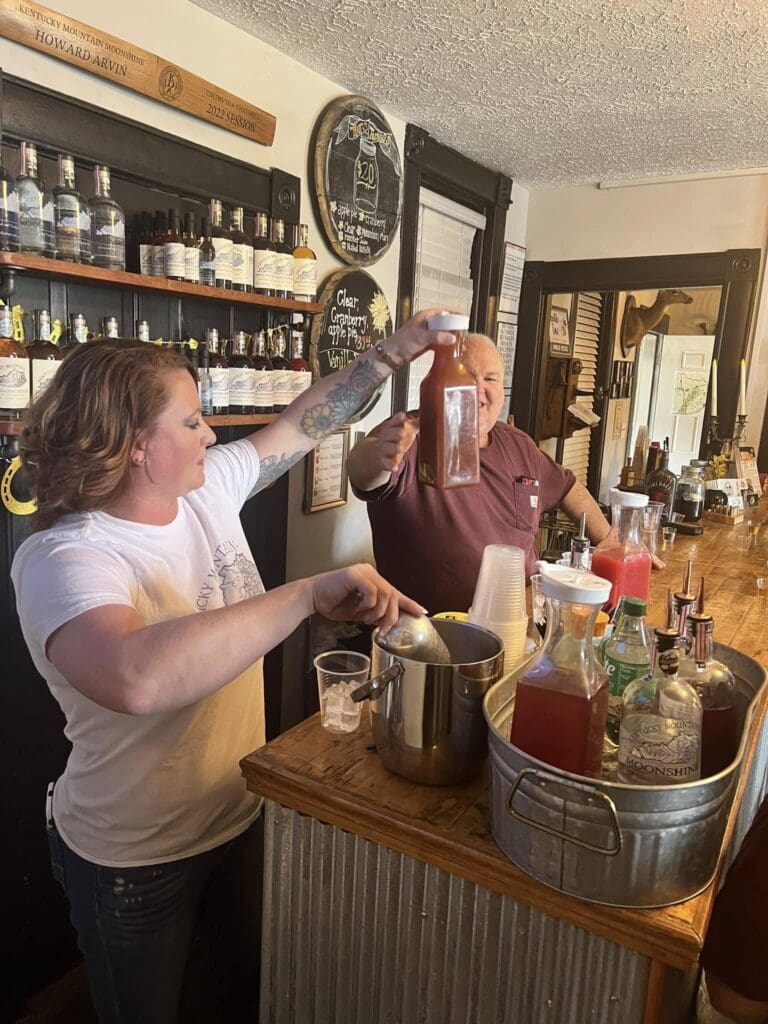 Bar counter scene: a woman pours from a large bottle into a metal tub while a man smiles nearby; shelves of liquor bottles line the back wall.