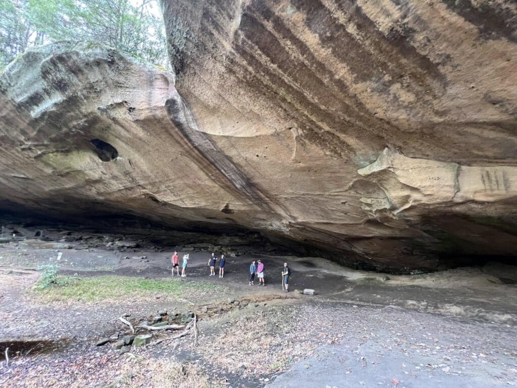 Group of people standing inside a large cave with layered rock ceiling and dirt floor.