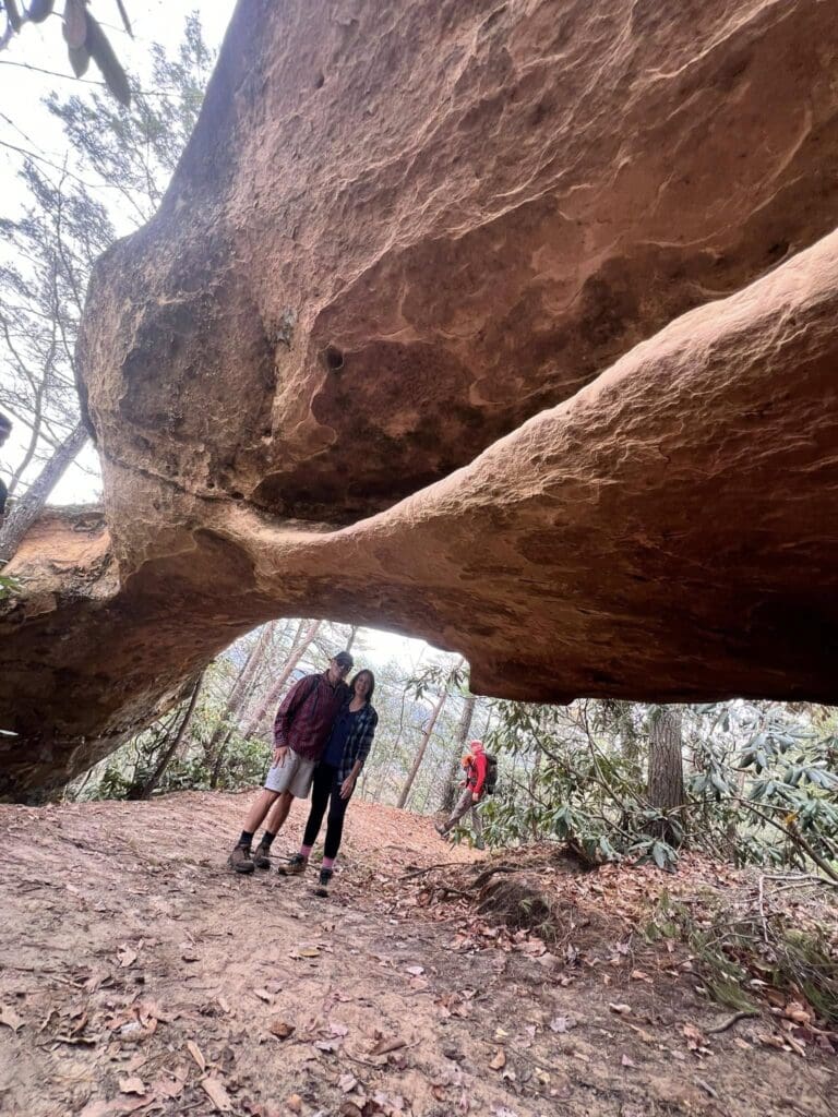 Group of hikers standing beneath a massive curved rock formation in a forest, fallen leaves on the ground.
