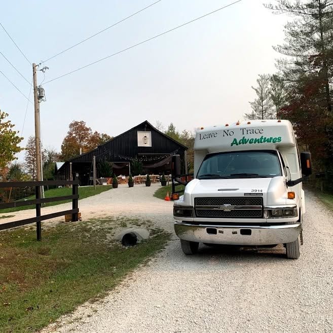 White shuttle van with 'Leave No Trace Adventures' parked on a gravel driveway in front of a rustic barn with string lights and outdoor seating nearby.