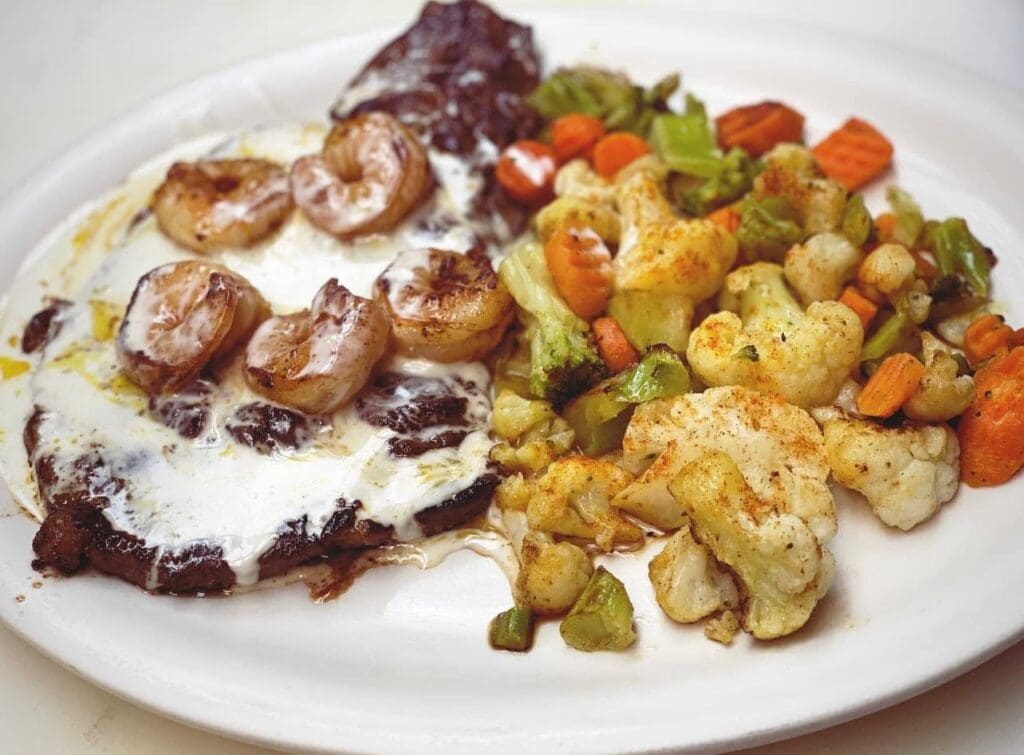 Steak with creamy white sauce topped with shrimp, served with roasted cauliflower and mixed vegetables on a white plate.