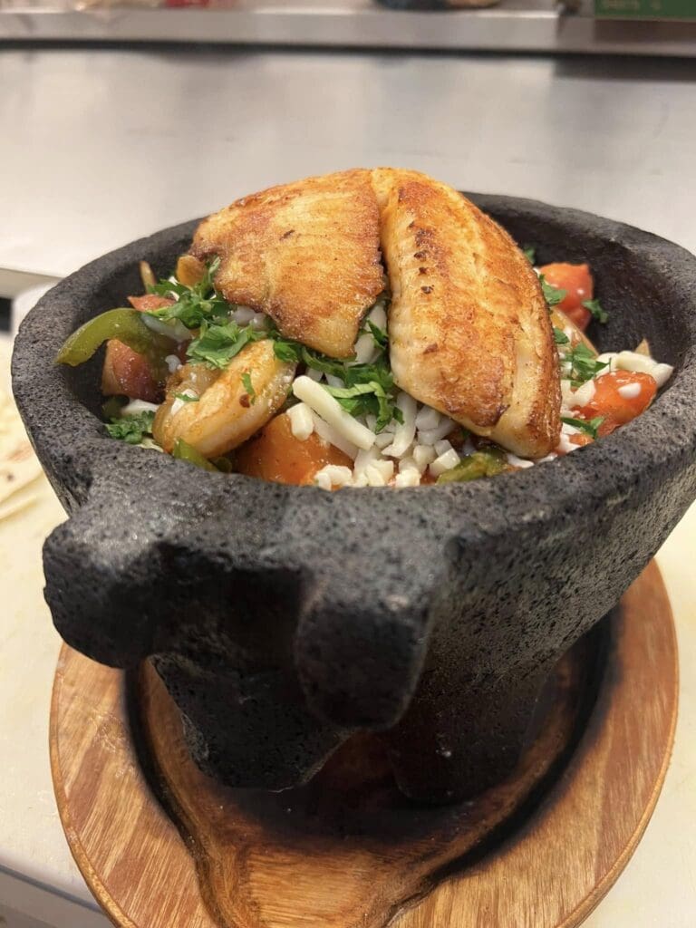 Two pan-seared salmon fillets rest on a bed of rice and chopped vegetables in a dark stone bowl (molcajete).
