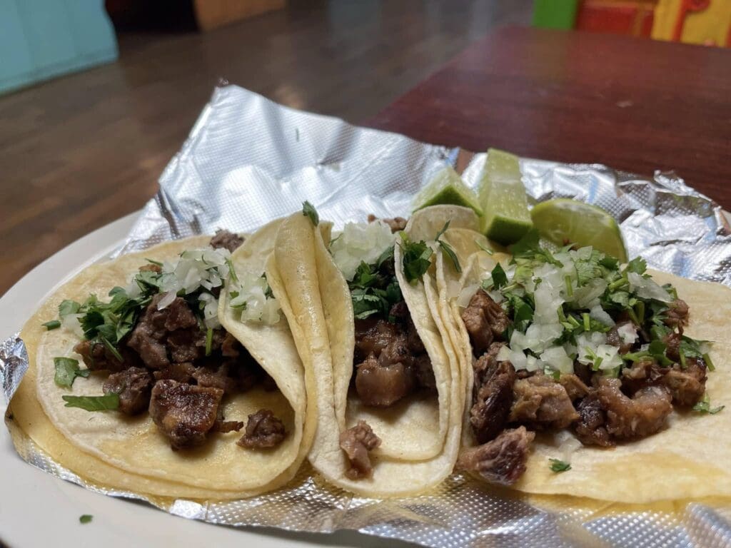 Three soft tortillas filled with grilled beef, chopped onions, cilantro, served on a foil-lined plate with lime wedges in the background.