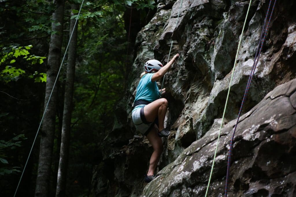 Climber wearing a helmet ascends a rocky cliff with safety ropes in a forest setting.