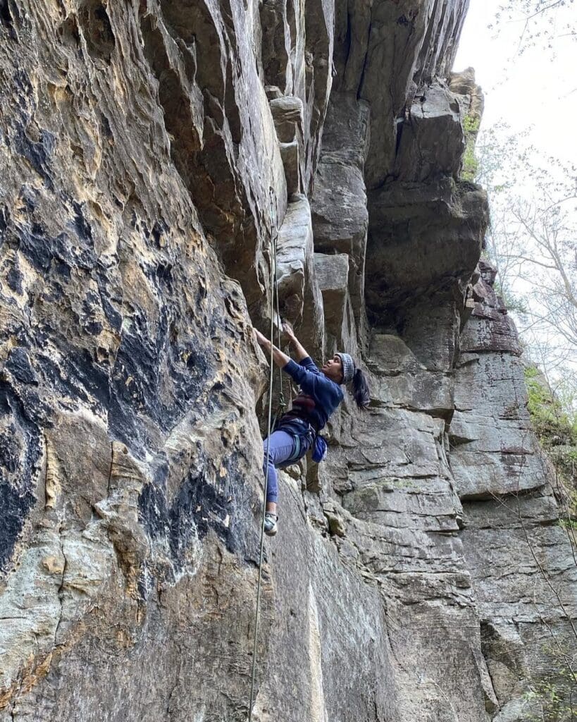 Climber wearing a headband ascends a rugged rock cliff with a safety rope.