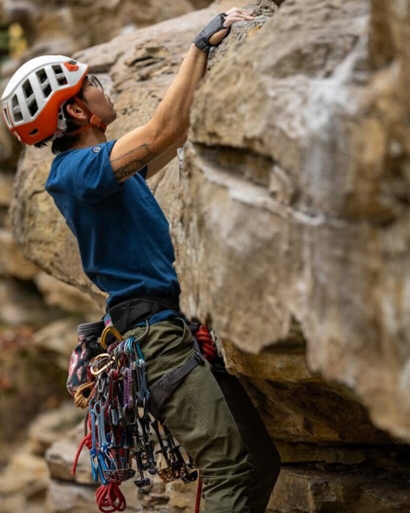 Climber in a blue shirt and orange helmet grips a rock face, equipped with a belt of carabiners and ropes.